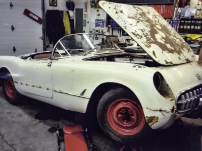 1954 Corvette side profile on lift showing original white paint and red wheels before restoration