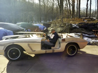 Front three-quarter of 1962 Corvette body in primer outdoors with autumn foliage behind