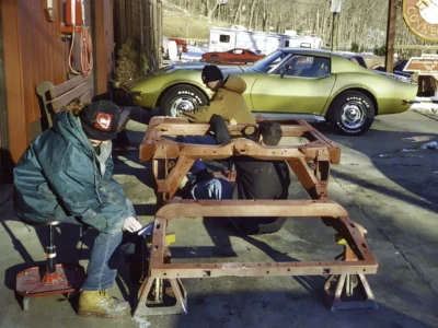 Technicians working on a 1968 Corvette frame mounted on a wooden support fixture.