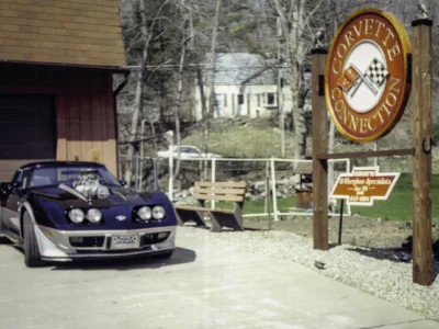 Remastered photo of a blue classic performance coupe parked near the Corvette Connection sign in Bolton, Connecticut, showing a front angle view and restored exterior details.