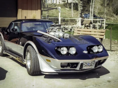 Remastered photo of a blue classic performance coupe front angle parked near the Corvette Connection sign in Bolton, Connecticut, showing custom front styling, wide tires, and restored exterior details.
