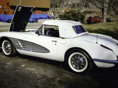 Remastered photo of a white 1959 Chevrolet Corvette convertible side view with hood open outside Corvette Connection, showing silver side coves, restored paint, and classic whitewall tires.
