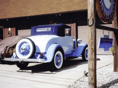 Remastered photo of a blue 1930s classic convertible side angle parked outside Corvette Connection in Bolton, Connecticut, showing whitewall tires, chrome trim, and restored paint.