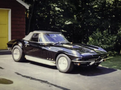 Remastered photo of a black 1965 Chevrolet Corvette coupe front three-quarter view outside Corvette Connection, showing the restored bodywork, chrome bumper detail, and classic mid-year Corvette styling.