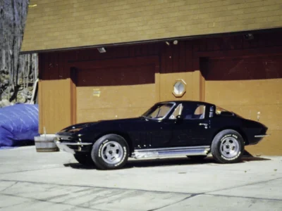 Remastered photo of a black 1965 Chevrolet Corvette coupe side profile parked outside Corvette Connection in Bolton, Connecticut, showing the fastback roofline, side exhaust area, and polished wheels.