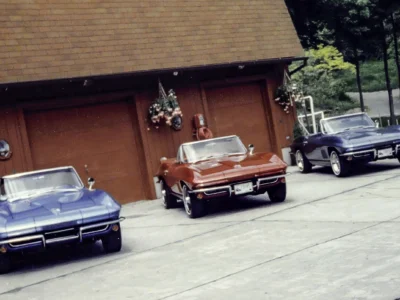 Remastered photo of a red 1965 Chevrolet Corvette convertible parked outside Corvette Connection, showing the front end, chrome bumper, black soft top, and restored exterior finish.