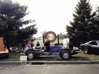 Remastered photo of a 1957 Chevrolet Corvette chassis restoration displayed at a Corvette Connection car show booth with classic vehicles in the background.