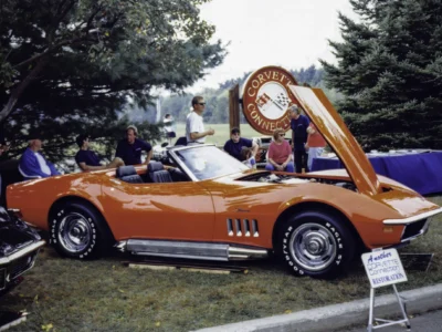 Remastered photo of a red 1965 Chevrolet Corvette convertible displayed at a local car show by Corvette Connection in Bolton, Connecticut, showing the side profile, chrome side pipes, and restored red paint.