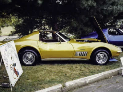 Remastered photo of a yellow 1966 Chevrolet Corvette convertible side profile with hood open at a car show, showing restored paint, chrome side trim, and classic Corvette styling.