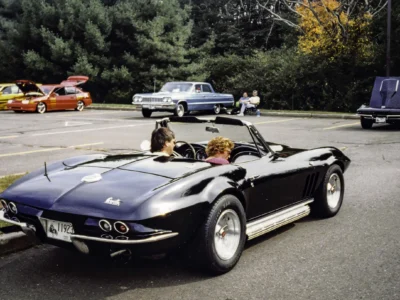 Remastered photo of a black 1965 Chevrolet Corvette convertible in a parking lot, showing passengers seated inside and classic mid-year Corvette styling.