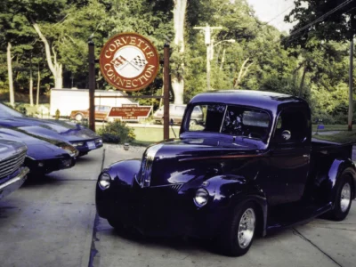 Remastered photo of a blue classic hot rod coupe parked near the Corvette Connection sign in Bolton, Connecticut, showing restored paint, rounded fenders, and custom street rod details.